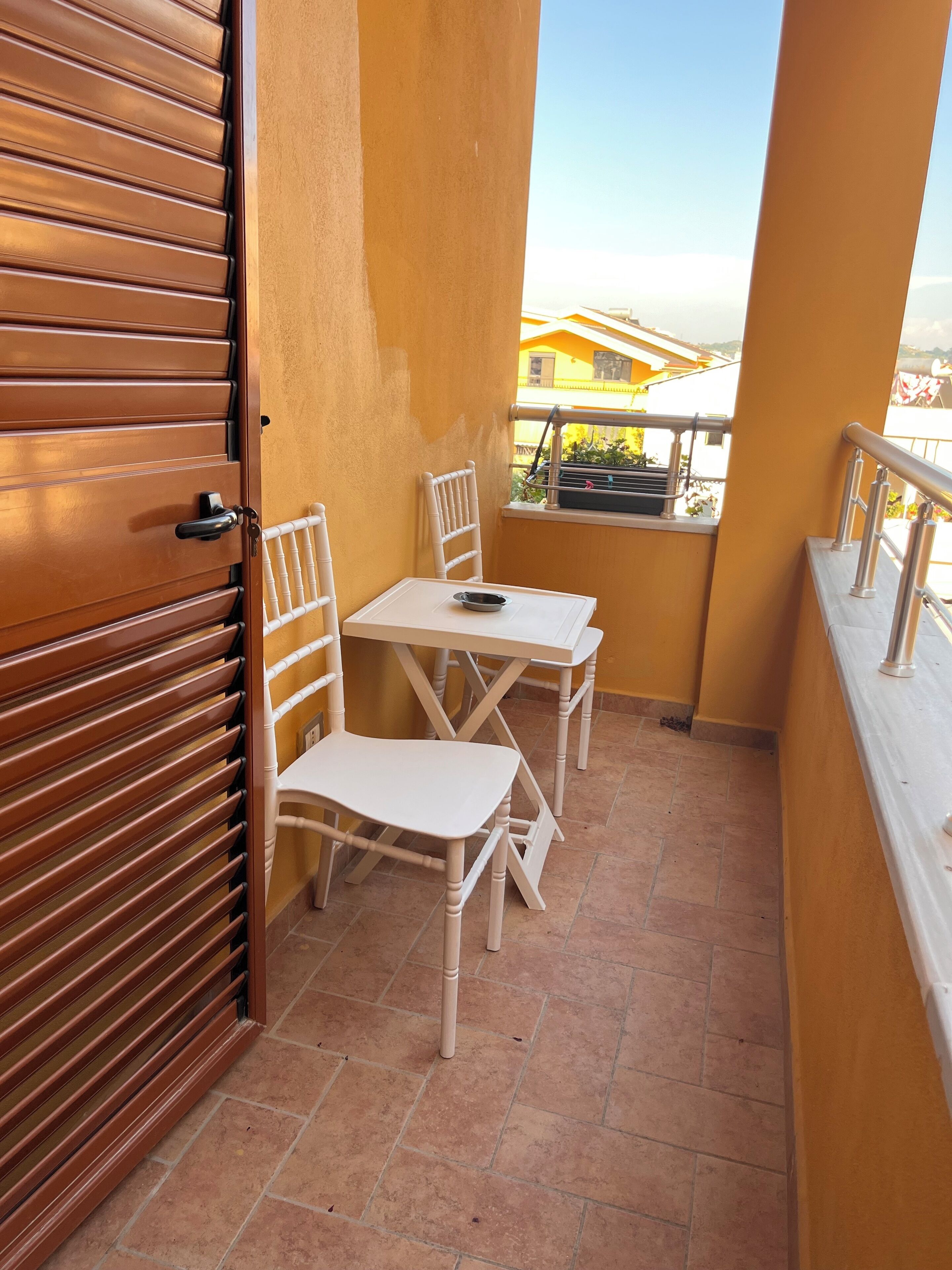 A balcony with a wooden door, white chairs, a small table, and a view of buildings in the background.