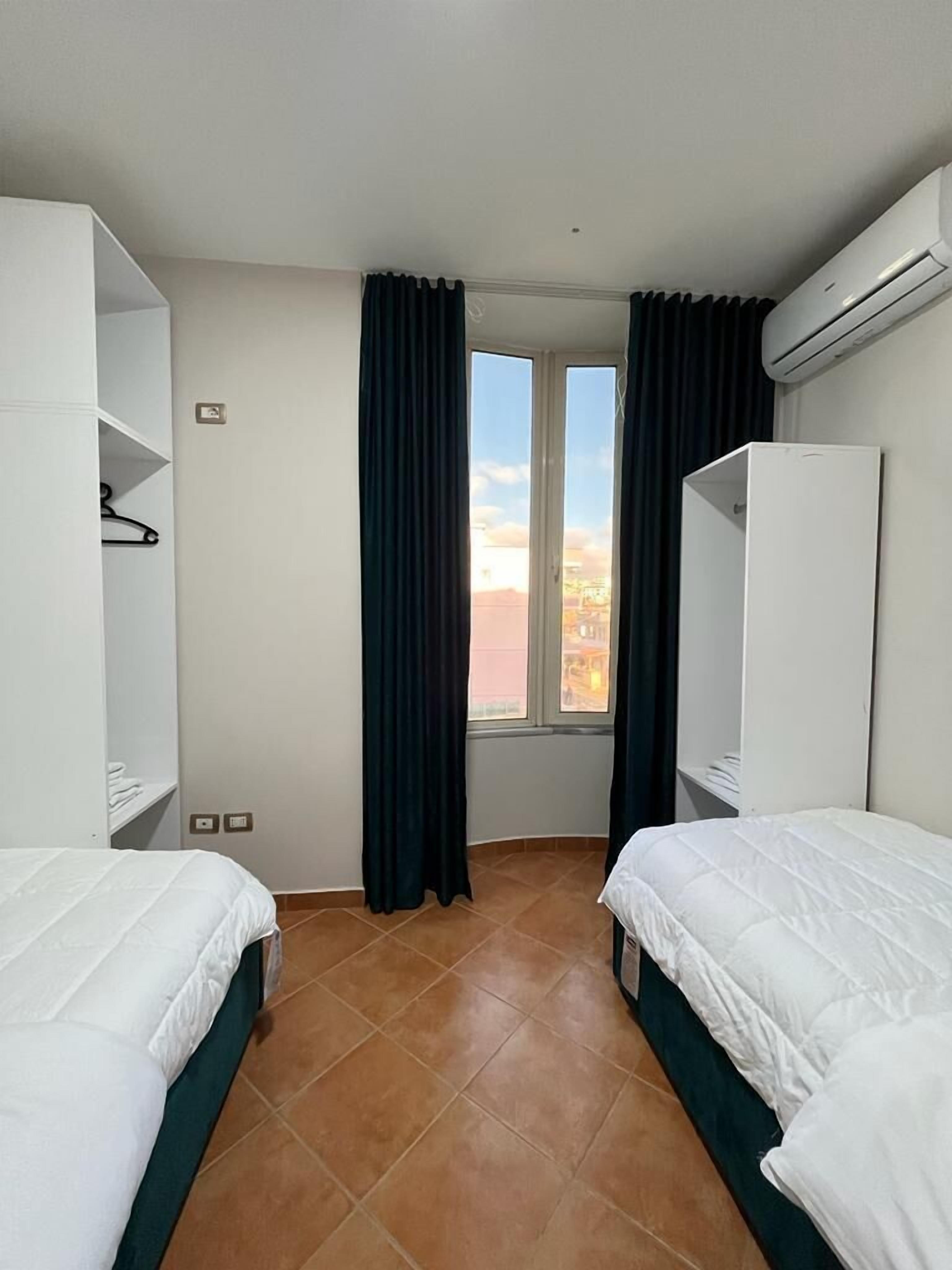 A hotel room with two beds, a wardrobe, a window with curtains, and an air conditioning unit.