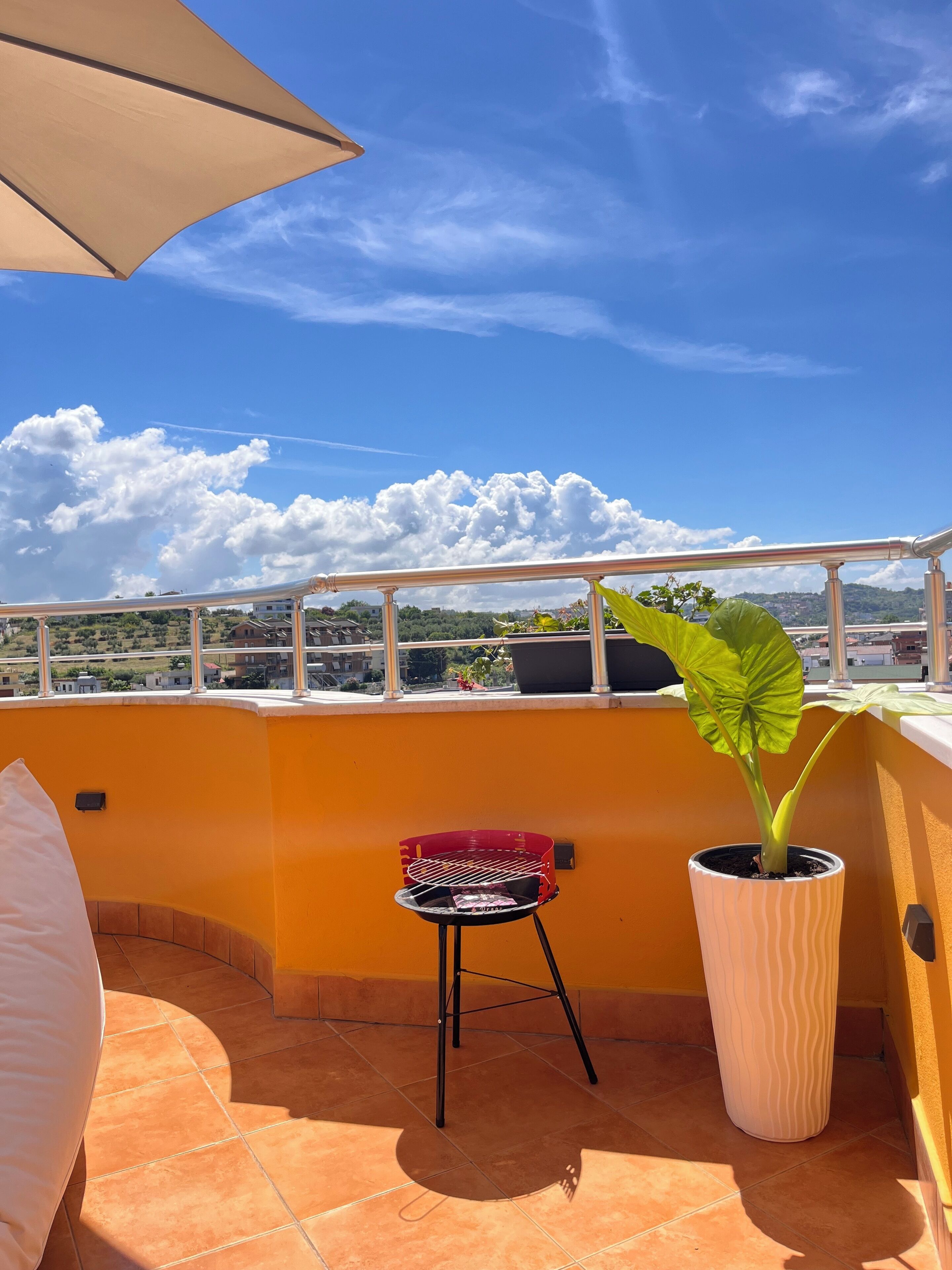 A rooftop terrace with a yellow wall, a red chair, a large potted plant, and a white umbrella.