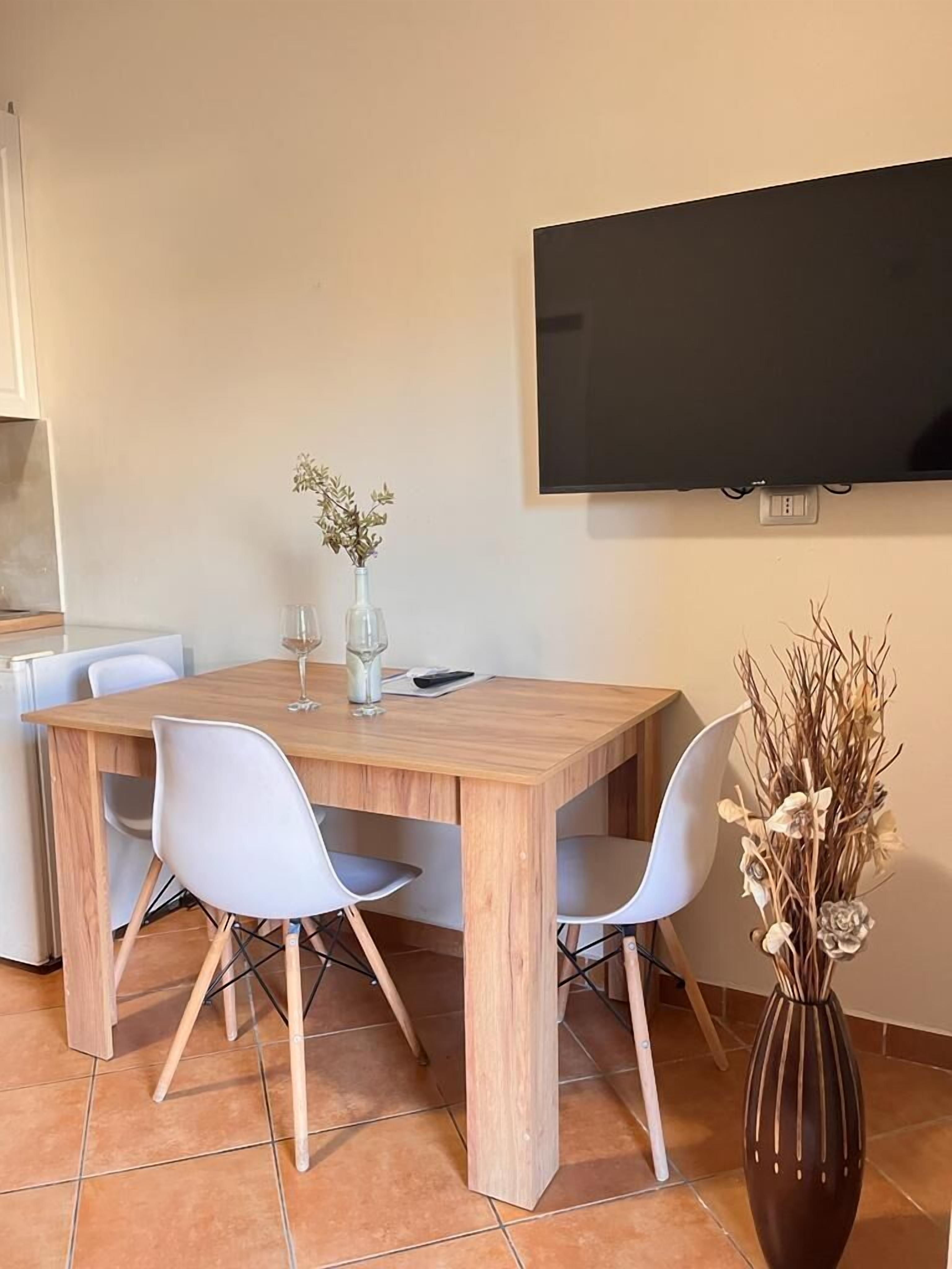A dining area with a wooden table, four white chairs, a vase with dried flowers, and a flat-screen TV mounted on the wall.