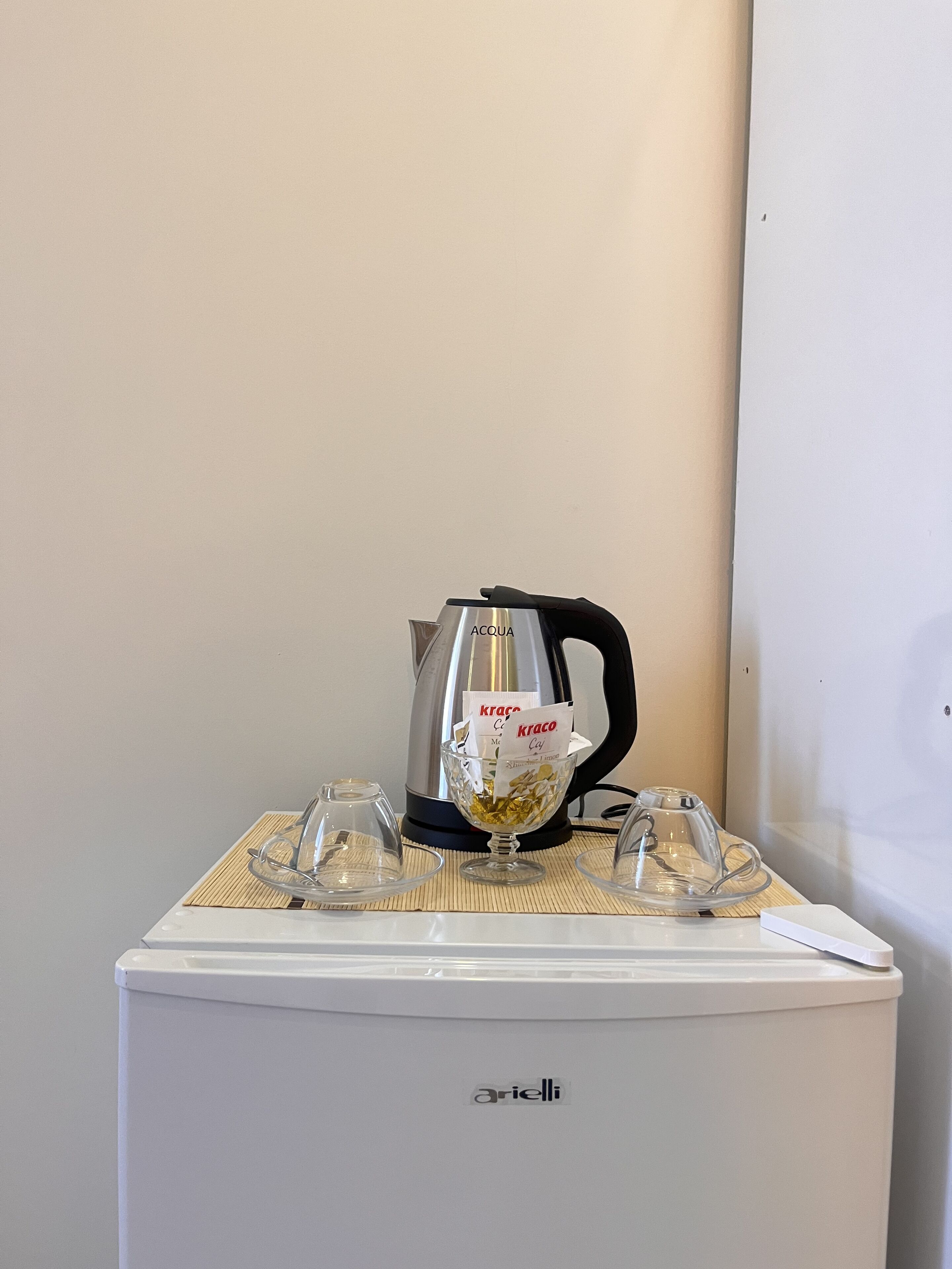 Una cocina compacta con un refrigerador blanco, un hervidor plateado y un recipiente de vidrio sobre una estera de bambú.