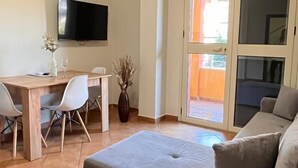 Un salon avec un canapé gris, une table à manger en bois avec des chaises, un téléviseur à écran plat fixé au mur et une porte-fenêtre coulissante donnant sur un espace extérieur.