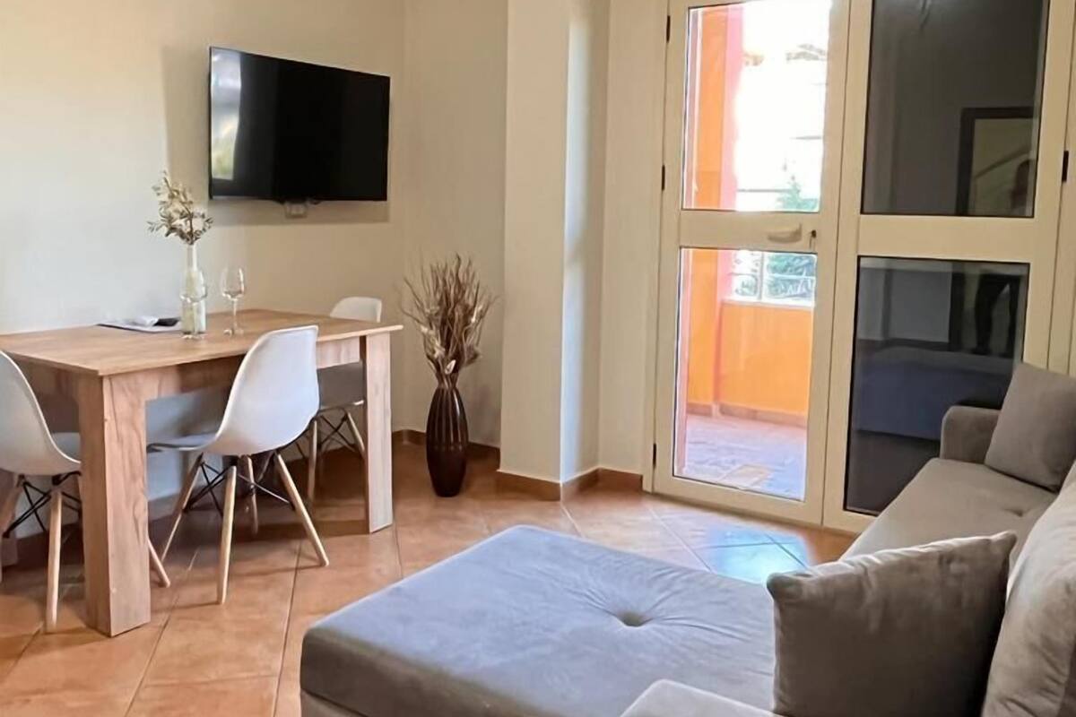 Un saló amb un sofà gris, una taula de dinar de fusta amb cadires, un televisor de pantalla plana encastat a la paret i una porta corredissa de vidre que dona a una zona exterior.
