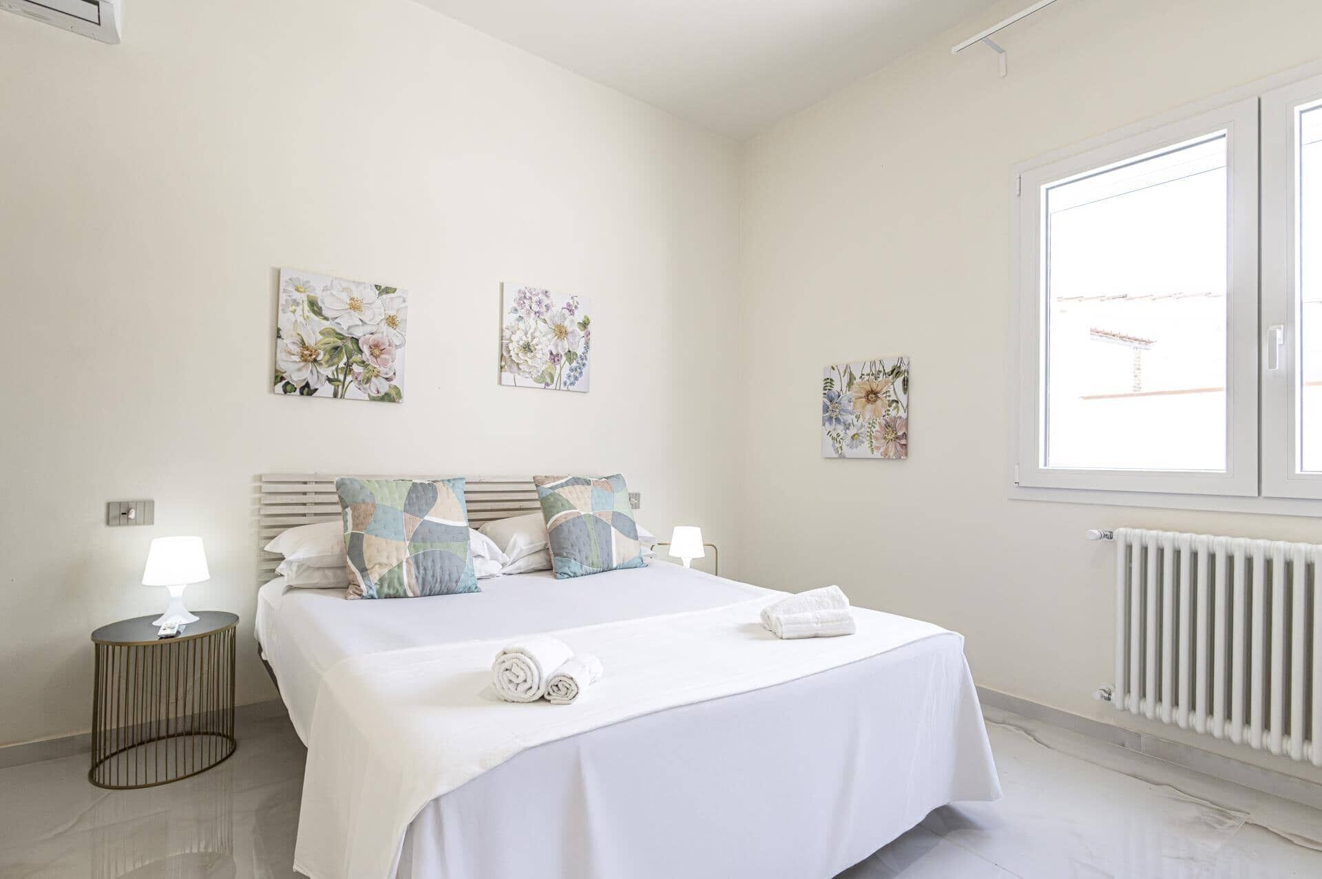 A bedroom with a bed, two wall-mounted flower paintings, a bedside table with a lamp, and a window with a view of a building.