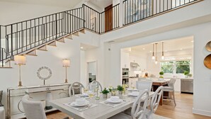 A spacious dining area with a high ceiling, a staircase with black railings, and a kitchen visible through an open doorway. - Lighthouse Dreams - Mesa Stunner Near the Beach (Santa Barbara)