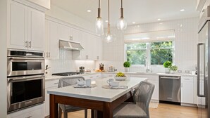 A modern kitchen with white cabinetry, stainless steel appliances, a central island with seating, pendant lighting, and a window providing natural light. - Lighthouse Dreams - Mesa Stunner Near the Beach (Santa Barbara)