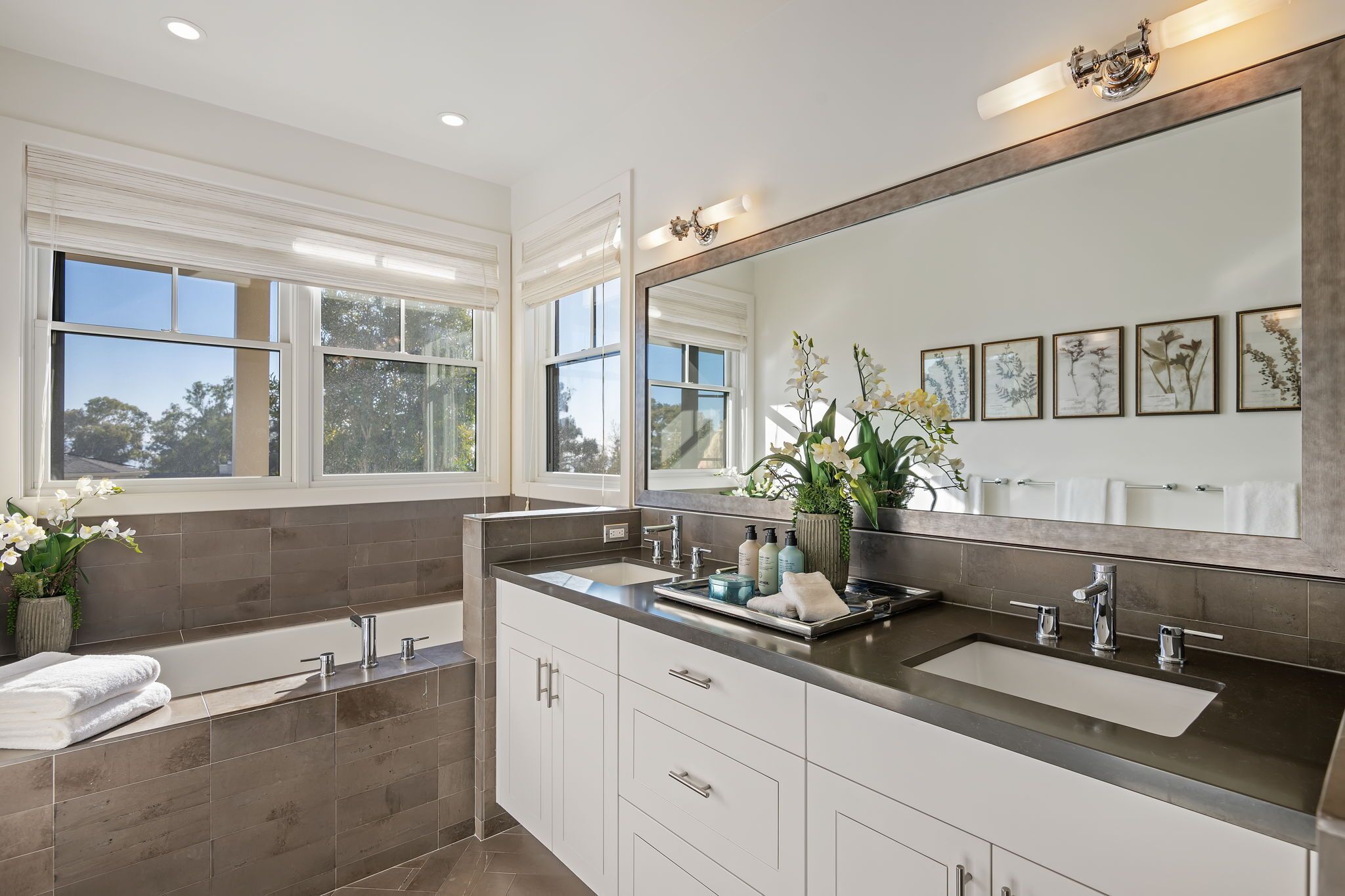 A modern bathroom with a large bathtub, a vanity with a sink, and a large mirror. There are framed pictures on the wall and a window with a view of trees.