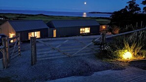 En landejendom med grusvej, der fører til en træport, en lille bygning med åben dør, og udsigt til havet i måneskin.