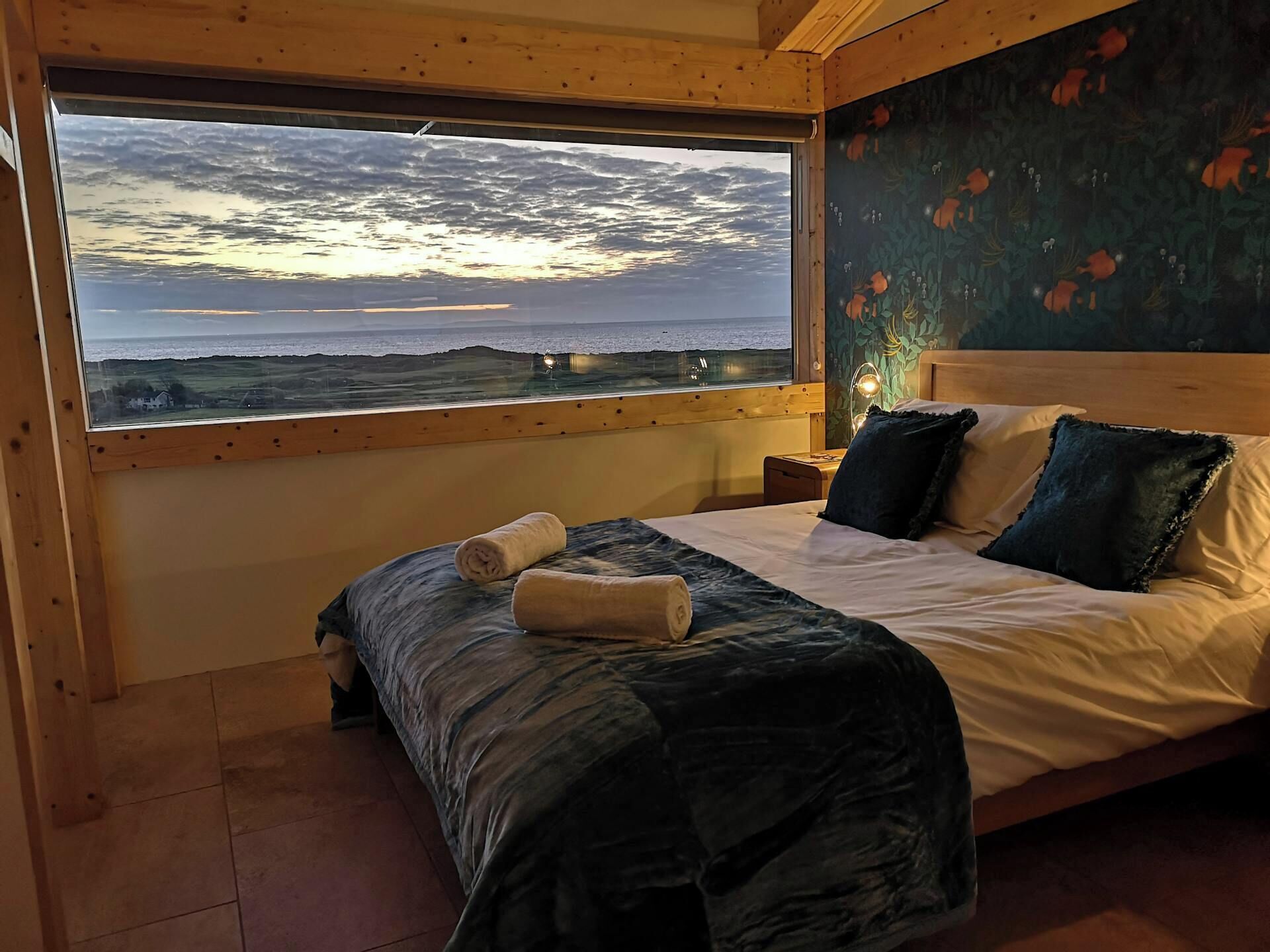 Ein Schlafzimmer mit großem Fenster, einem Bett mit Kissen, einem Nachttisch und einer Wand mit floralem Muster.