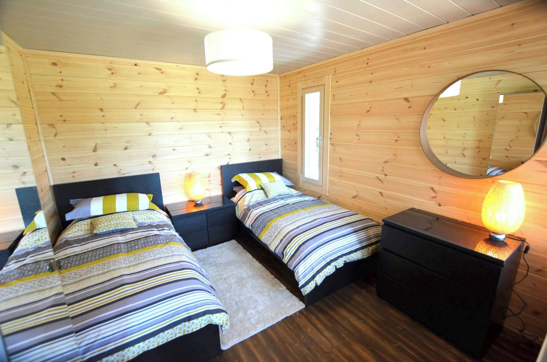 En stuga med träpanel, inredd med två sängar, en spegel och ett nattduksbord med en lampa.