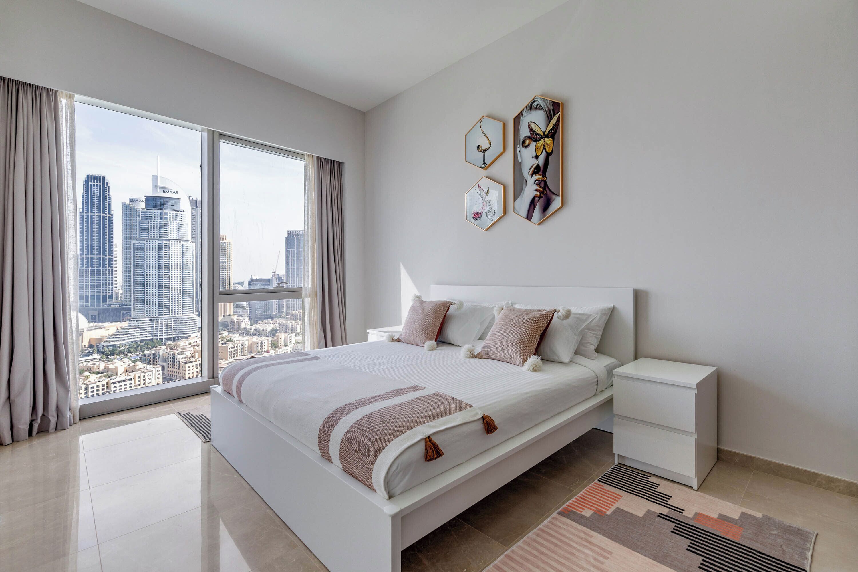 Une chambre moderne avec un grand lit, une table de chevet et une vue sur le paysage urbain grâce à de grandes fenêtres du sol au plafond.