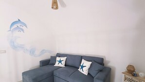 Ein graues Ecksofa mit zwei sternförmigen Kissen, Wandgemälde von Delfinen, eine gewebte Pendelleuchte und ein Holztisch mit Korb und Figur.