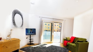 Una sala de estar moderna con suelo de madera, un sofá verde, un tapete negro, un mueble de madera y un televisor de pantalla plana sobre un pedestal.