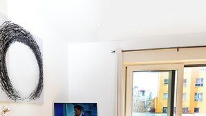 Una sala moderna con un televisor de pantalla plana, piso de madera, una alfombra oscura, un mueble de madera y un ventanal con vista a edificios.