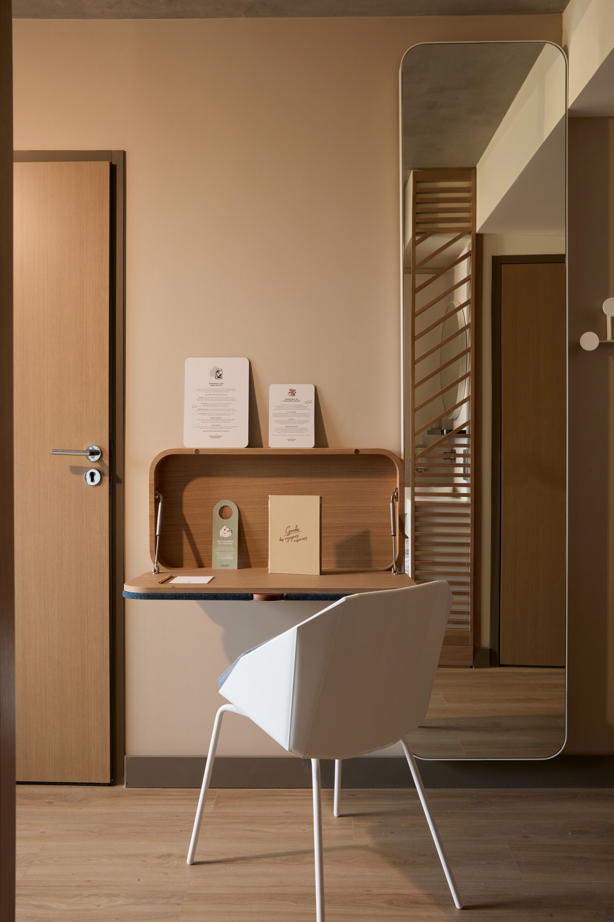 Une chambre d’hôtel moderne avec un bureau en bois, une chaise blanche et un miroir qui reflète un escalier.
