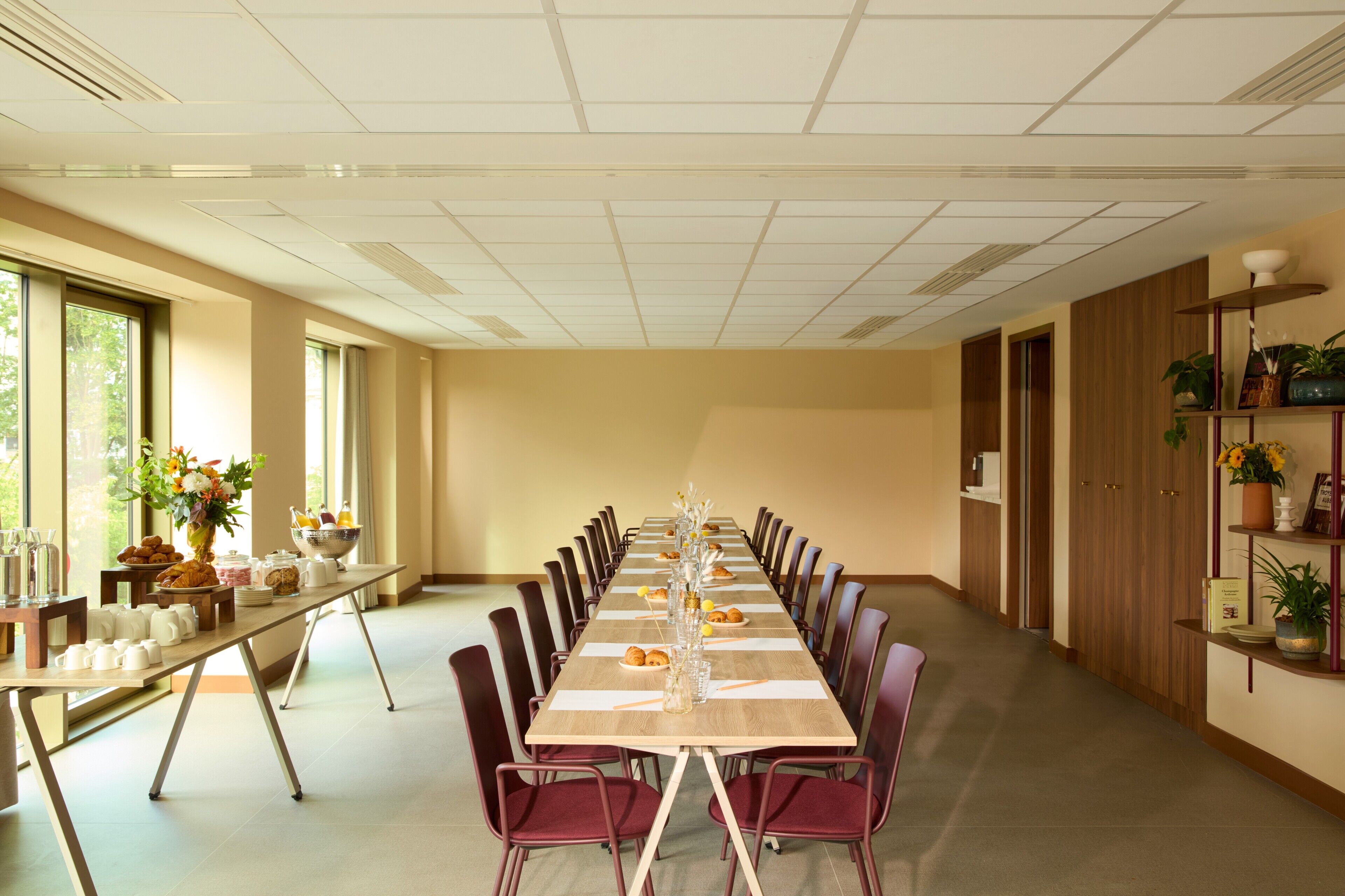 Une longue table de conférence entourée de chaises, un vase rempli de fleurs et une table de buffet garnie de plats et de boissons.
