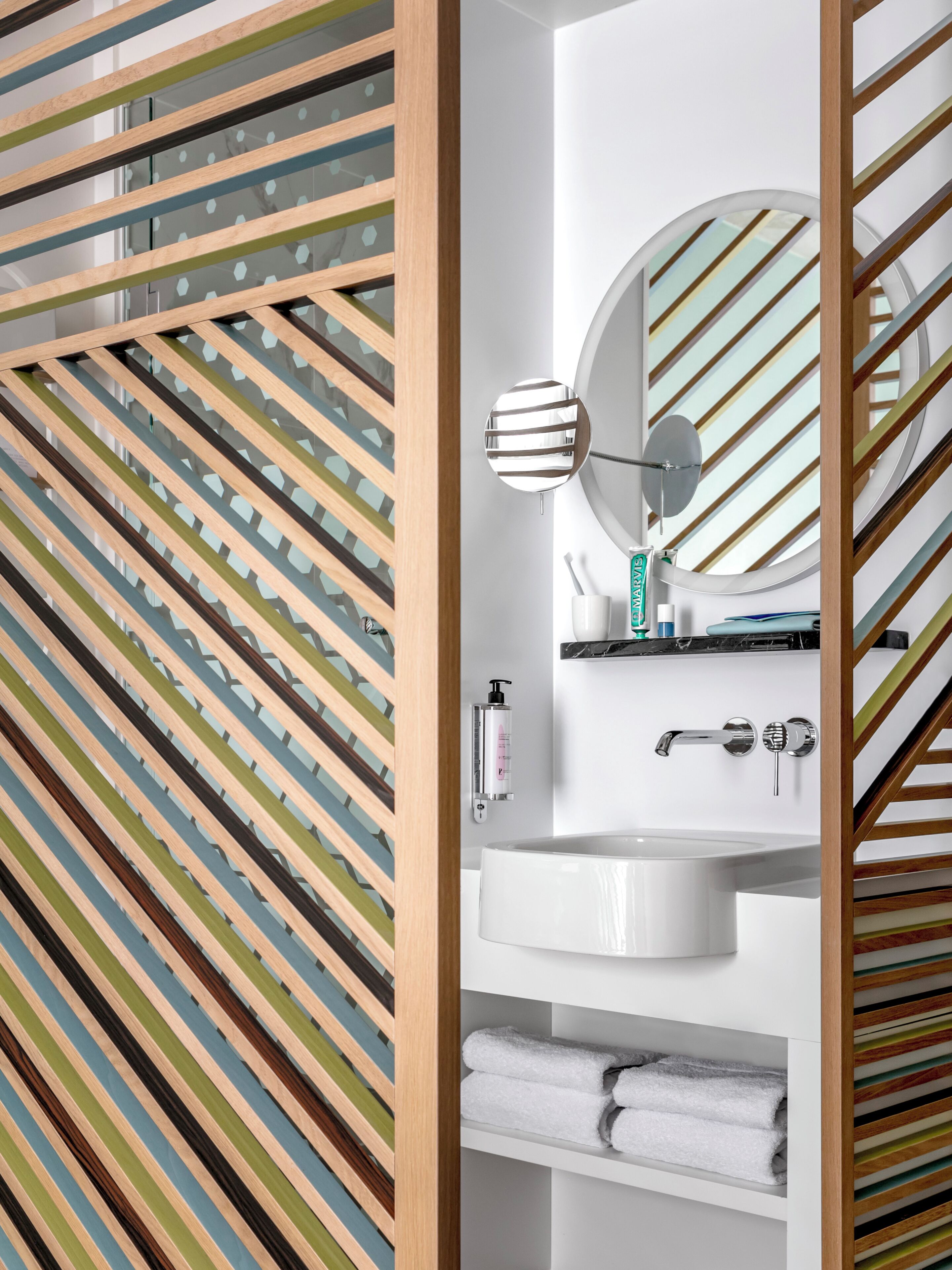 A modern bathroom with a striped wooden panel design, a round mirror, a white sink, and neatly folded towels.