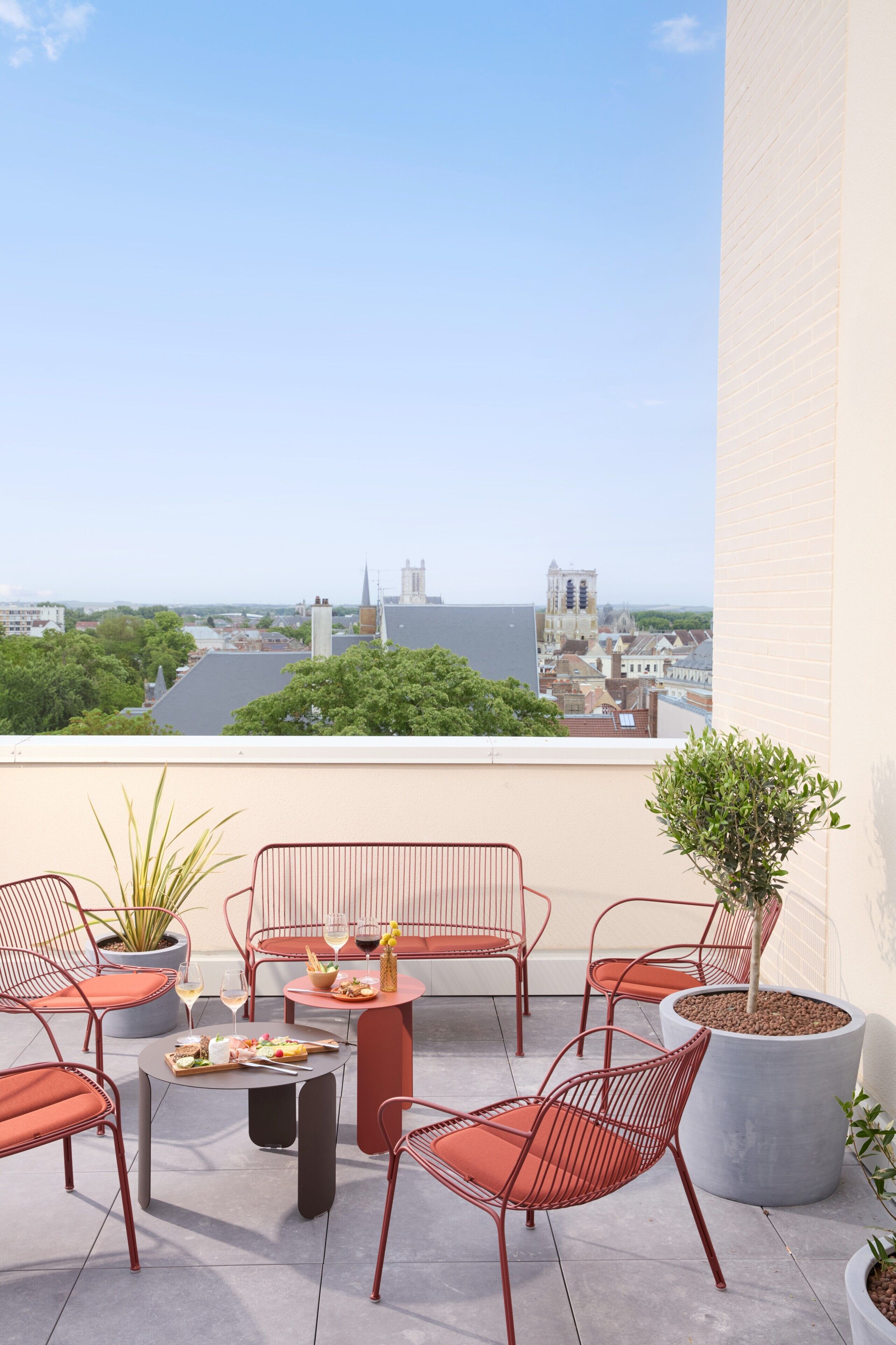 Una terraza en la azotea con sillas metálicas rojas, una mesa con comida y bebidas, plantas en macetas y vistas de la ciudad.