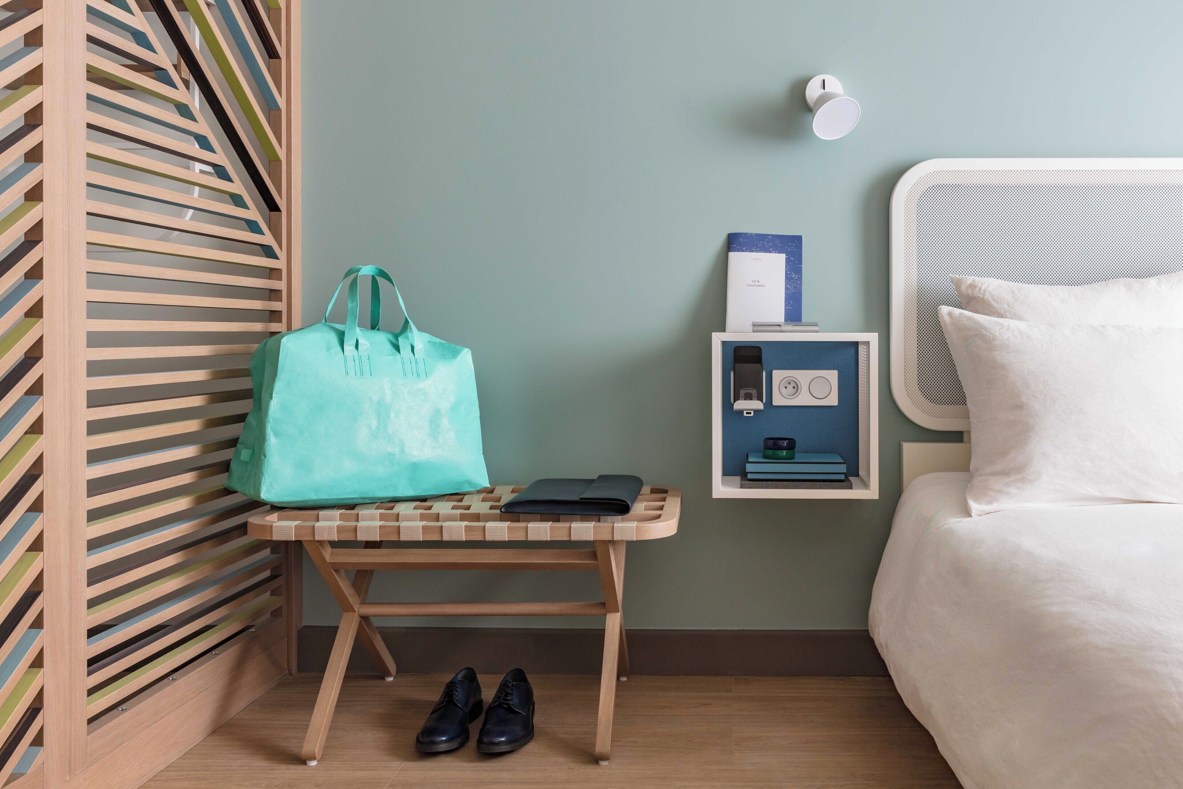 A hotel room with a bed, a nightstand with a lamp, a wall-mounted shelf with a phone, and a bench with a bag and shoes.