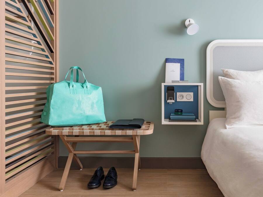Habitación de hotel con cama, mesita de noche con lámpara, estante en la pared con un teléfono, y un banco con bolso y zapatos.