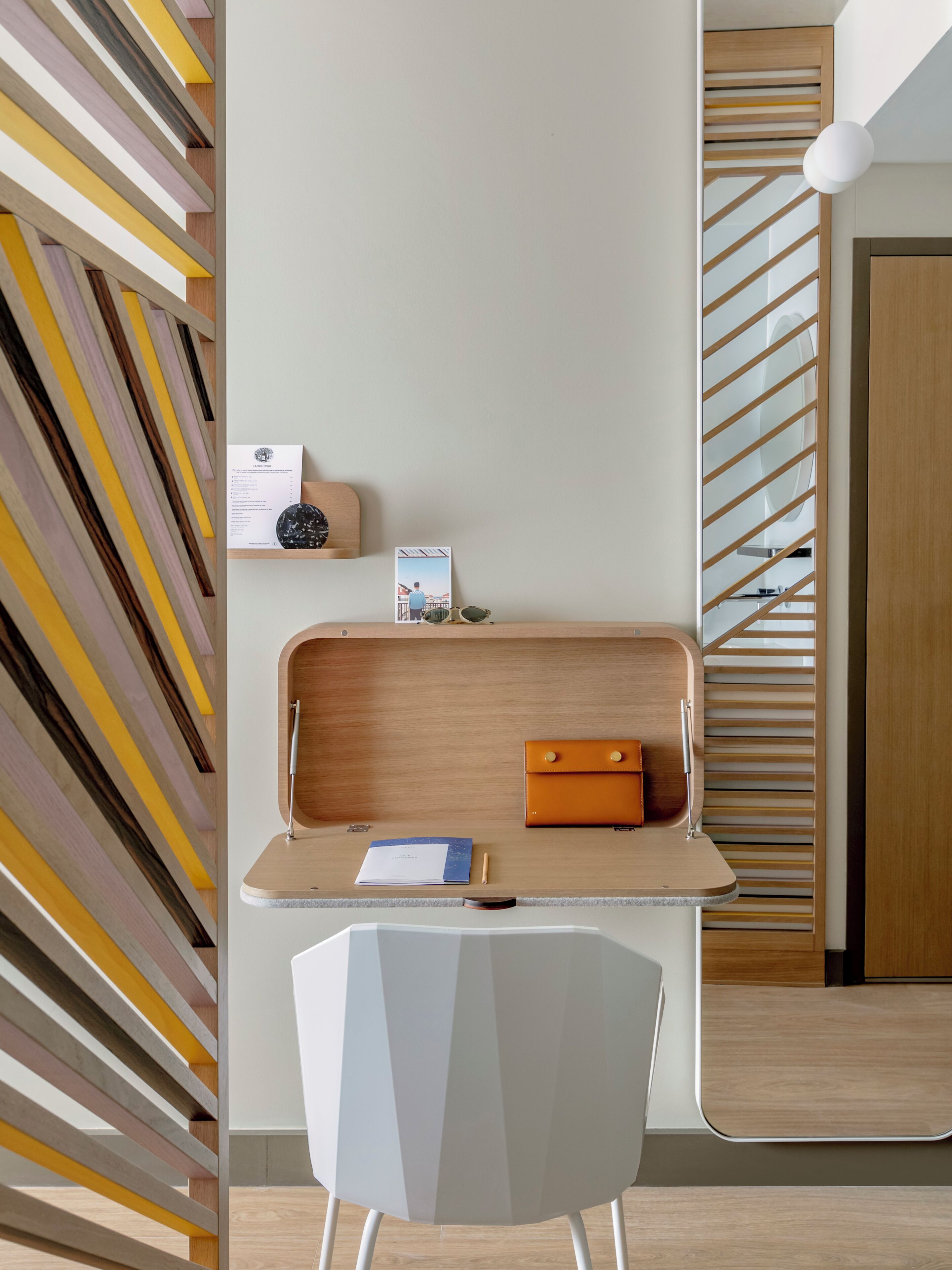 Een moderne, minimalistische kamer met een wand met een geometrisch patroon, een houten bureau met een laptop, een notitieboekje en een pen, en een witte stoel.