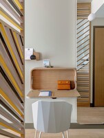 Una habitación moderna y minimalista con un muro de diseño geométrico, un escritorio de madera con una computadora portátil, un cuaderno y un bolígrafo, y una silla blanca.