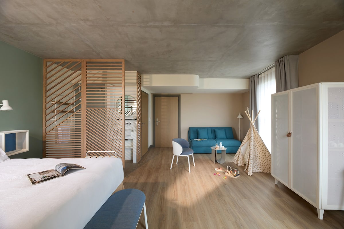 Een moderne hotelkamer met een bed, een blauwe bank, een klein tafeltje, een witte kast en een houten scheidingswand.