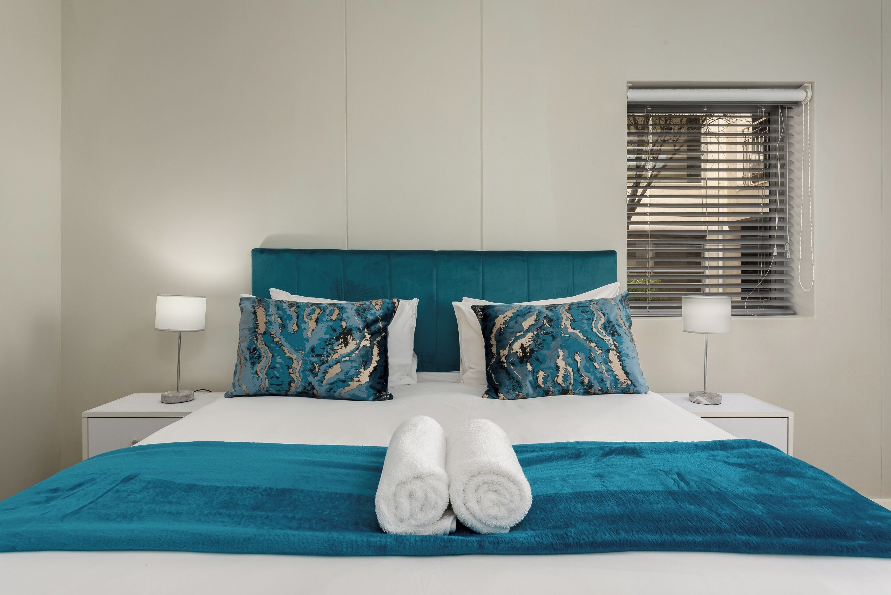 Una cama bien hecha con un cabecero color turquesa, dos almohadas a juego y un par de toallas enrolladas.