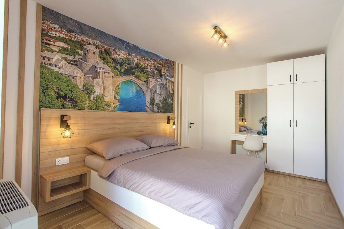 Une chambre à coucher comprenant un lit, une tête de lit en bois, une table de chevet, une commode, un miroir, une armoire et un tableau représentant une ville côtière, accroché au mur.
