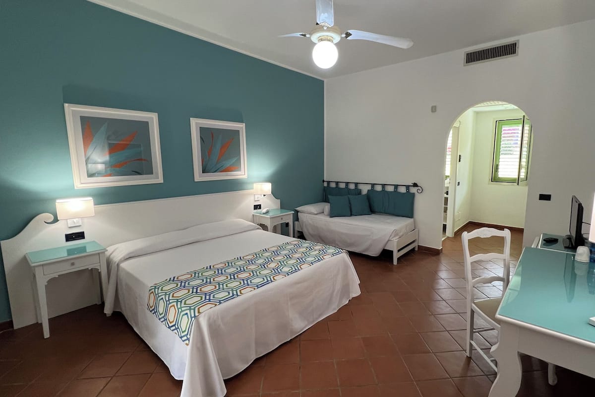 Ein Hotelzimmer mit zwei Betten, einem Schreibtisch mit Stuhl, einem Deckenventilator und Bildern an den Wänden.