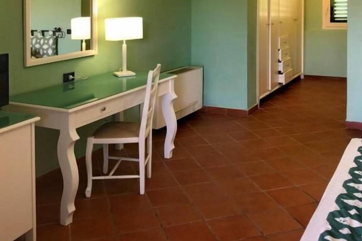 Ein Zimmer mit Schreibtisch, Stuhl und Spiegel. Ein Kleiderschrank und ein Fenster mit Jalousien sind ebenfalls vorhanden.