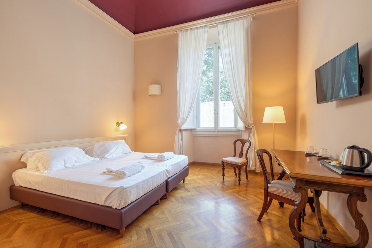 A hotel room with a bed, a chair, a table with a kettle, and a TV on the wall.