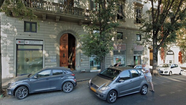 Un edificio con una amplia entrada en arco, un cartel que dice 'Monte dei Paschi di Siena' y dos coches estacionados en la calle.