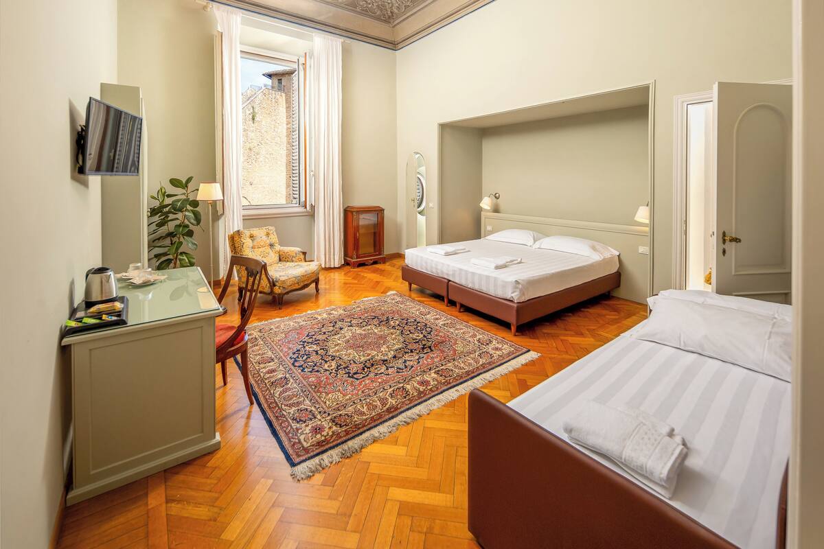 A hotel room with a large bed, a smaller bed, a desk with a chair, a mirror, and a window with curtains.