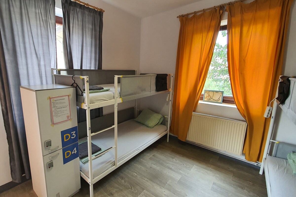 A dormitory room with bunk beds, a desk with a computer, and a radiator.