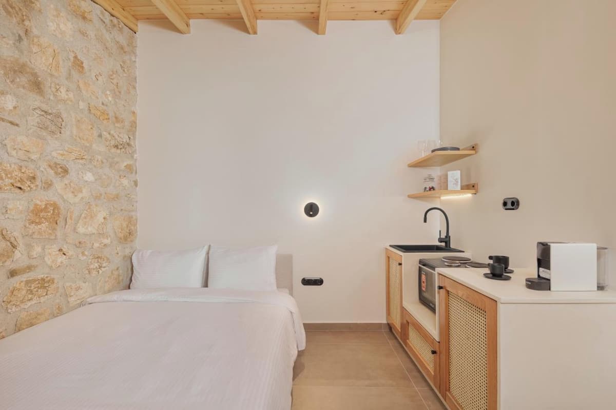 Quarto com parede de pedra, uma cama, uma pequena cozinha com pia e máquina de café, e uma prateleira com objetos.