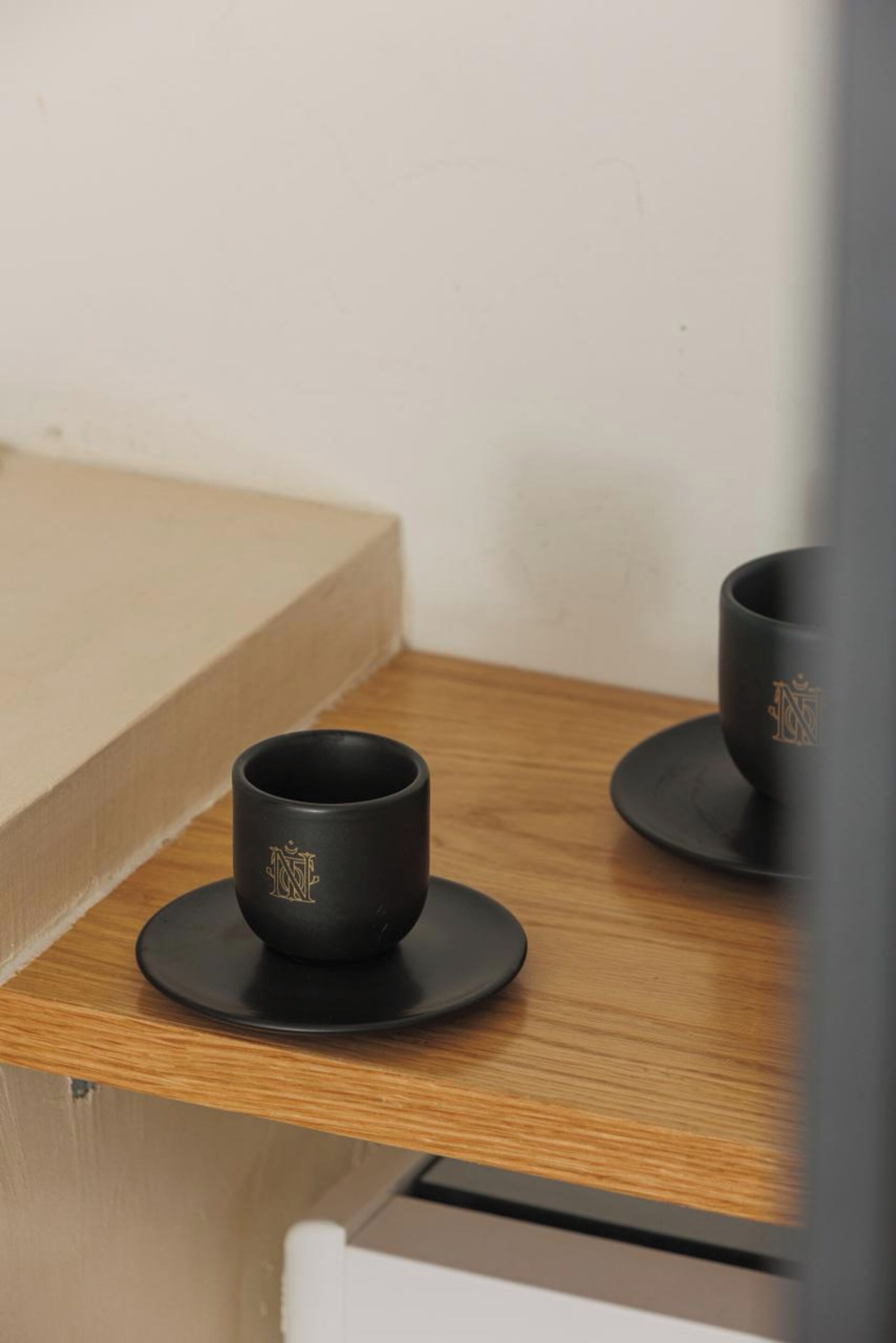 Une table en bois sur laquelle reposent deux tasses noires ornées d’emblèmes dorés, posées sur des soucoupes.