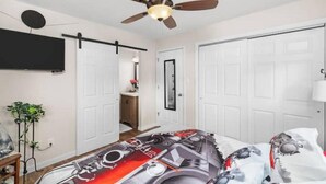 A bedroom with a ceiling fan, a bed with a mechanical-themed bedspread, a TV mounted on the wall, and white doors.