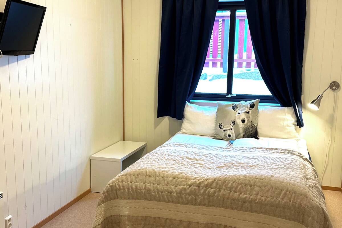 A bedroom with a bed, a nightstand, a flat-screen TV mounted on the wall, and a window with curtains.
