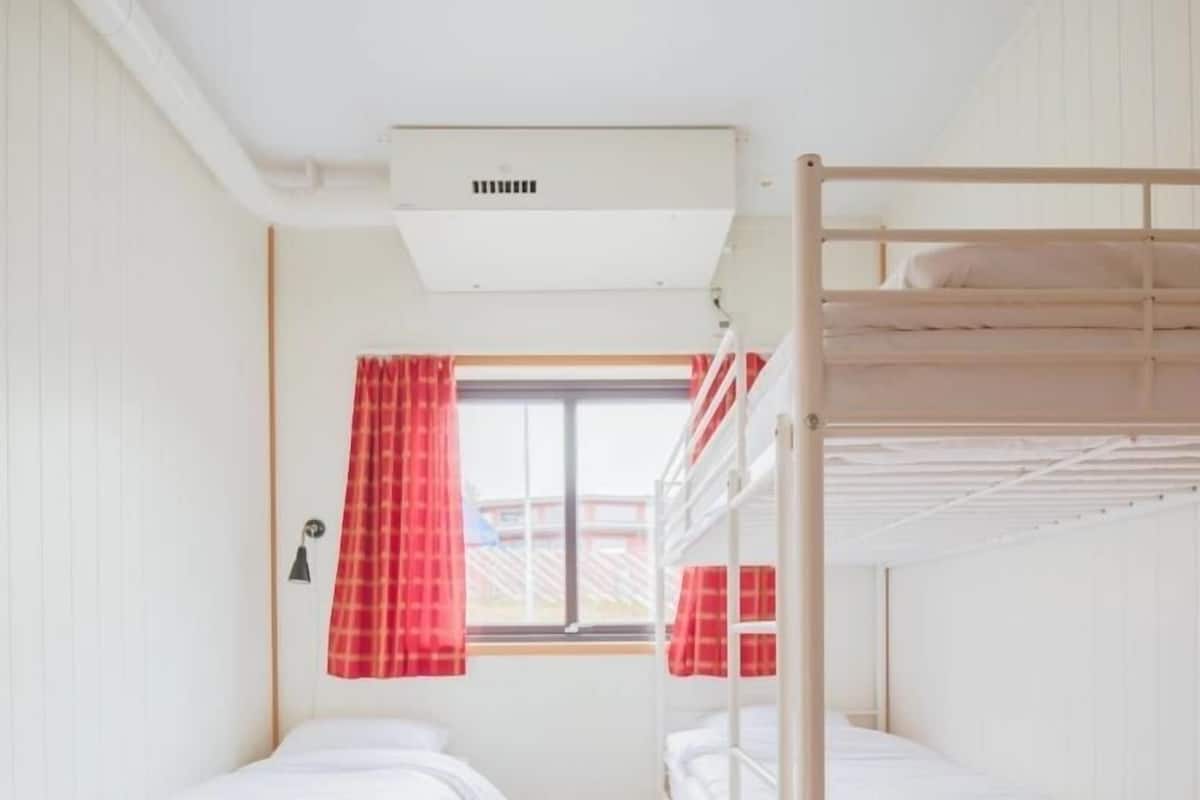 A dormitory room with bunk beds, a window with red curtains, and a ceiling vent.