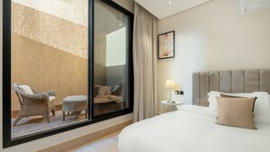 A hotel room with a large bed, a nightstand, a lamp, a wicker chair, a small table, and a view through a sliding glass door. - Qurtoba big apartment all yours (Riyadh)