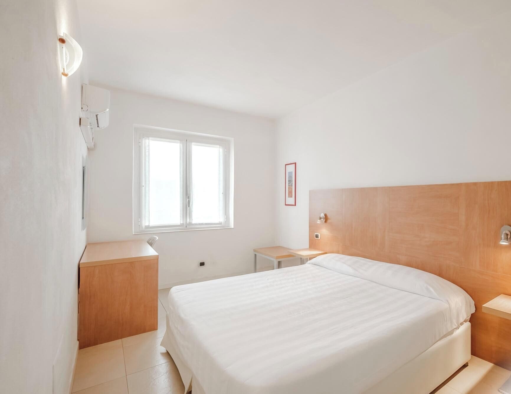 A hotel room with a large bed, bedside tables, a window, and a wall-mounted light fixture.