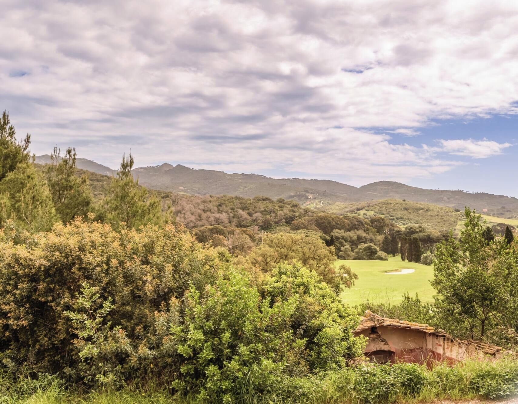 Paisaje rural con un campo de golf, vegetación densa y colinas al fondo.