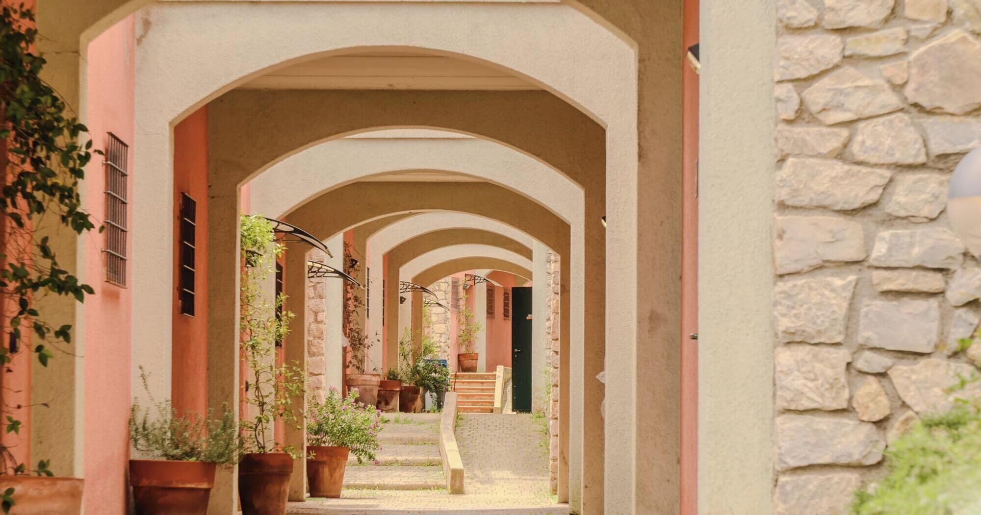 Une ruelle étroite, bordée d’arches, agrémentée de plantes en pot et d’un mur de pierre.