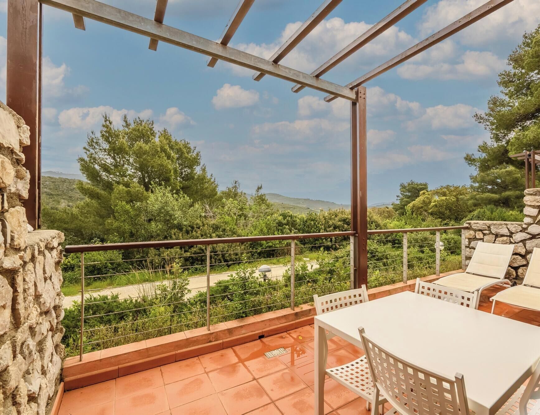 Un balcón con mesa y sillas blancas, con vistas a un paisaje exuberante con árboles y cielo despejado.