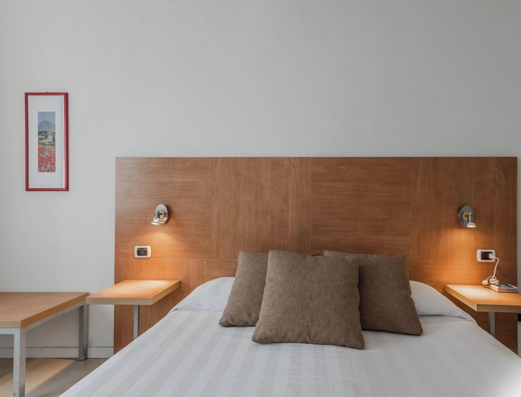 A hotel room with a wooden headboard, two bedside tables, a bed with white bedding, and a framed picture on the wall.