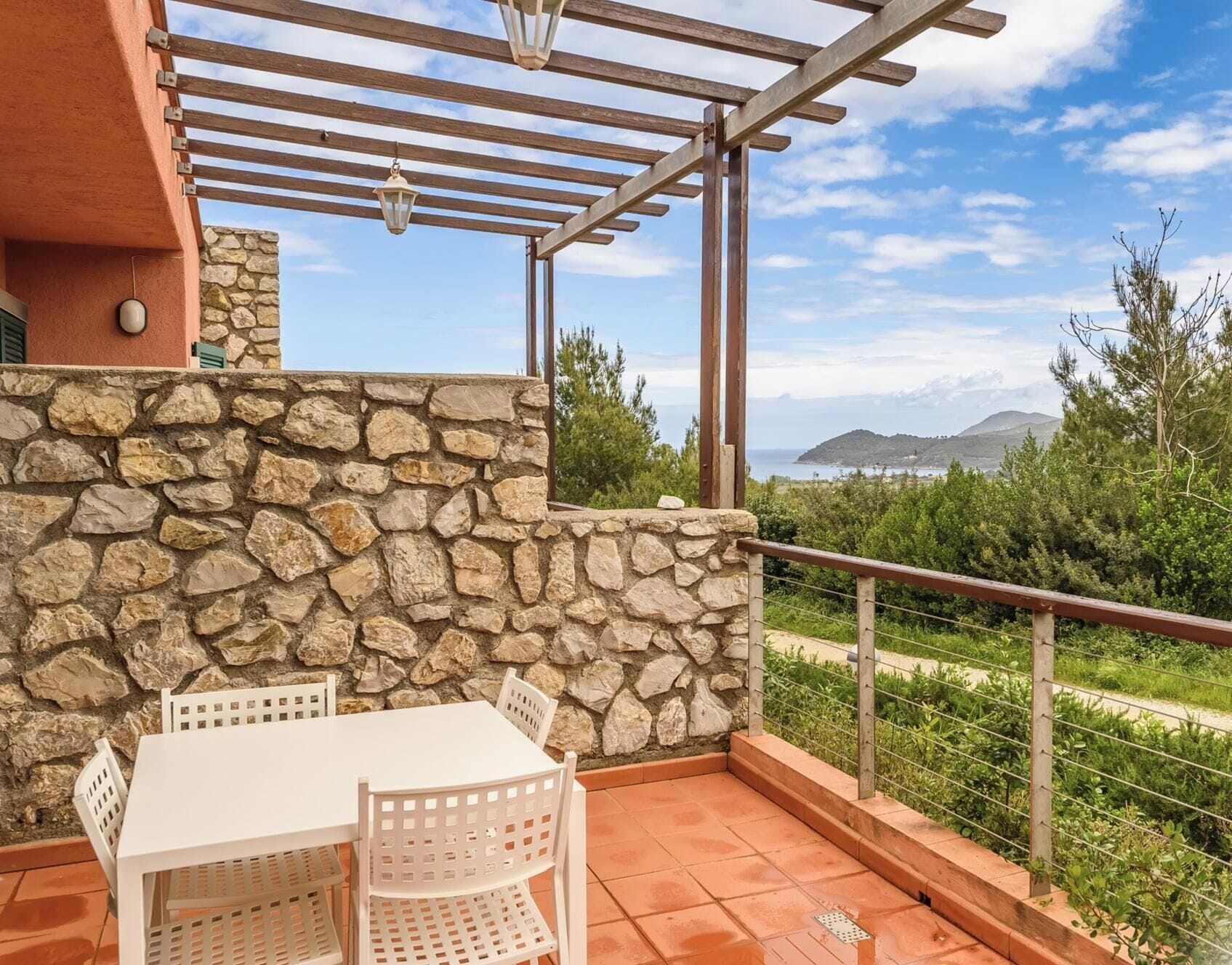A patio with a stone wall, a white table, and chairs, overlooking a coastal landscape.