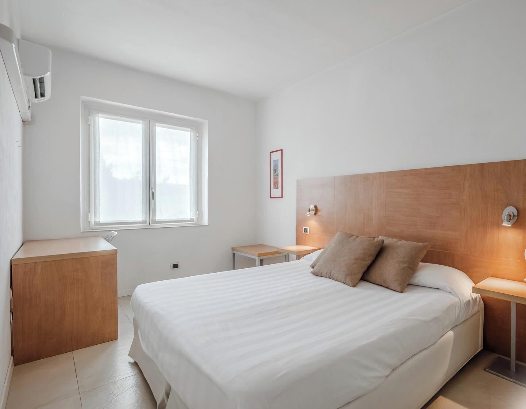 A hotel room with a large bed, bedside tables, a window, and a wall-mounted air conditioning unit.
