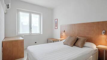 A hotel room with a large bed, bedside tables, a window, and a wall-mounted air conditioning unit.