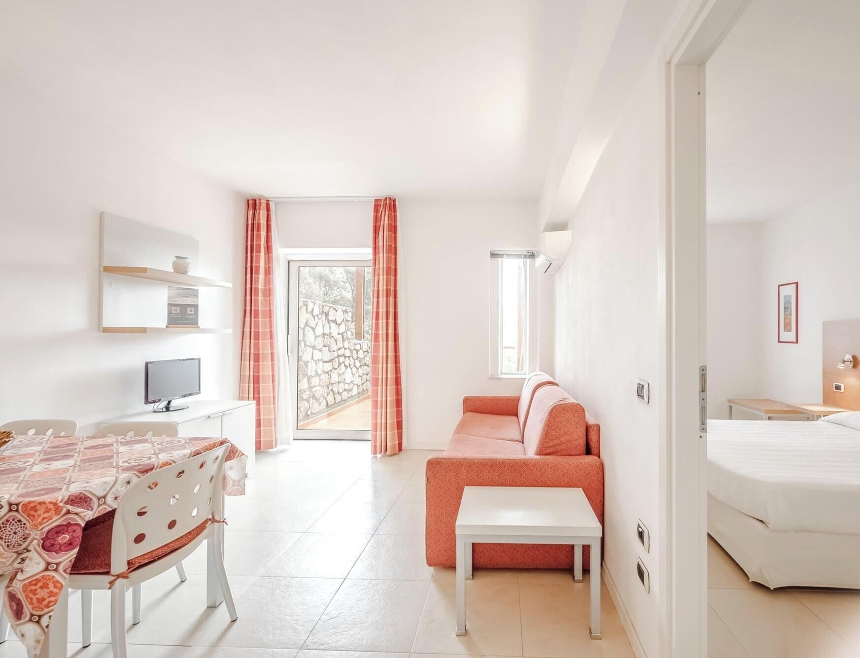 Une chambre d’hôtel moderne équipée d’un lit, d’un canapé, d’une table à manger avec des chaises, d’une télévision et d’un balcon agrémenté d’un mur en pierre.